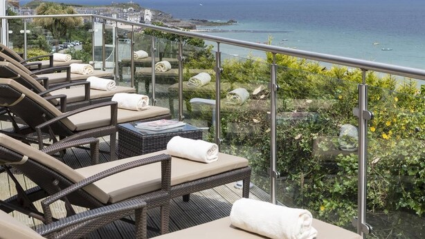 A balcony with wicker lounge chairs and a glass railing overlooking a coastal view.