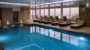An indoor swimming pool with clear blue water, surrounded by lounge chairs and large windows offering a view of the outdoors.