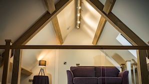 Ein Zimmer mit schrägem Dach, Holzbalken, einem violetten Sofa, einer Stehlampe und einem Koffer.
