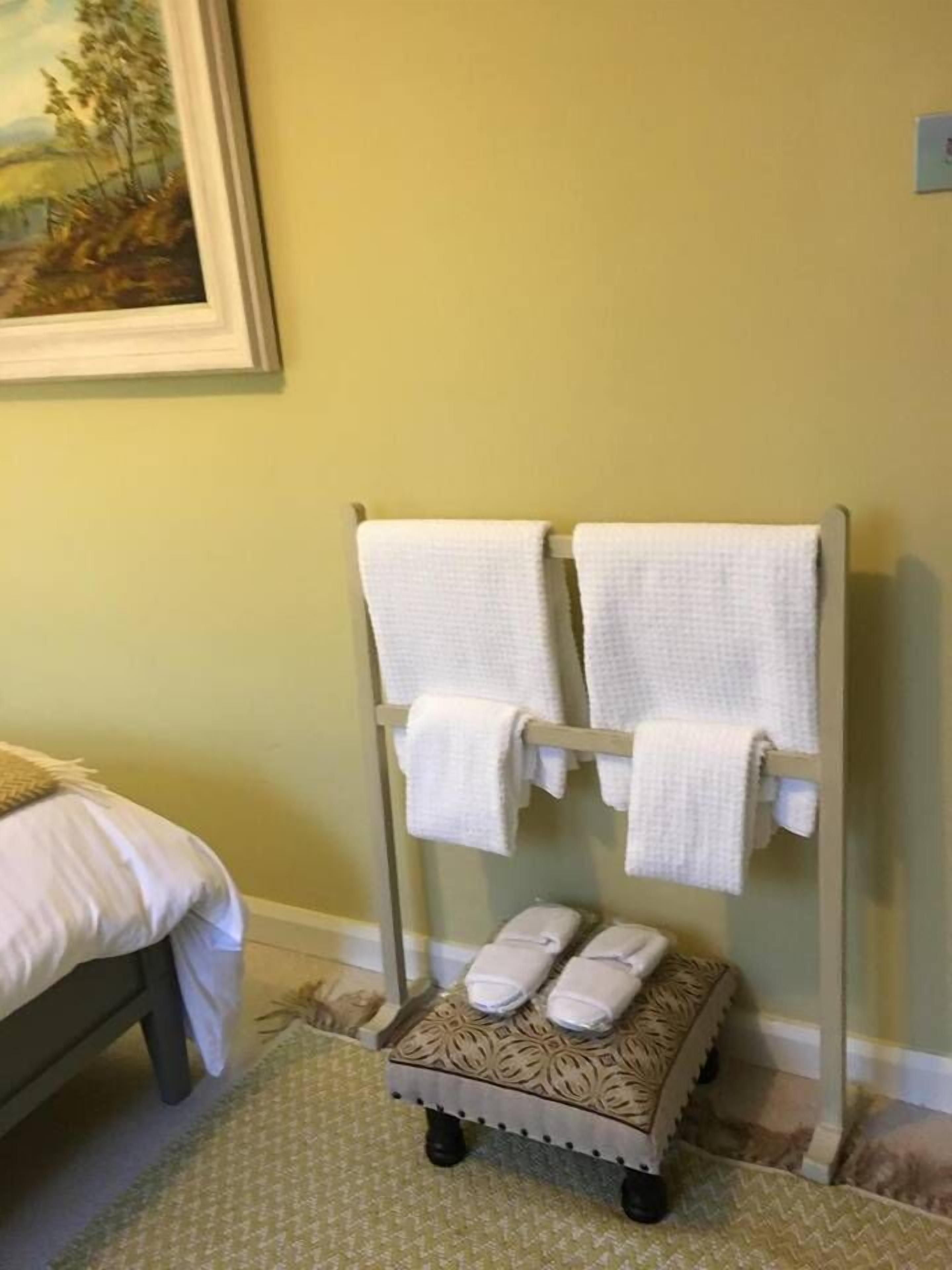 A bathroom with a towel rack holding two towels, a small table with folded towels, and a bed with a white bedspread.