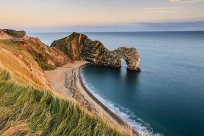 Paisagem costeira com um arco natural de rocha, uma praia arenosa e falésias gramadas.