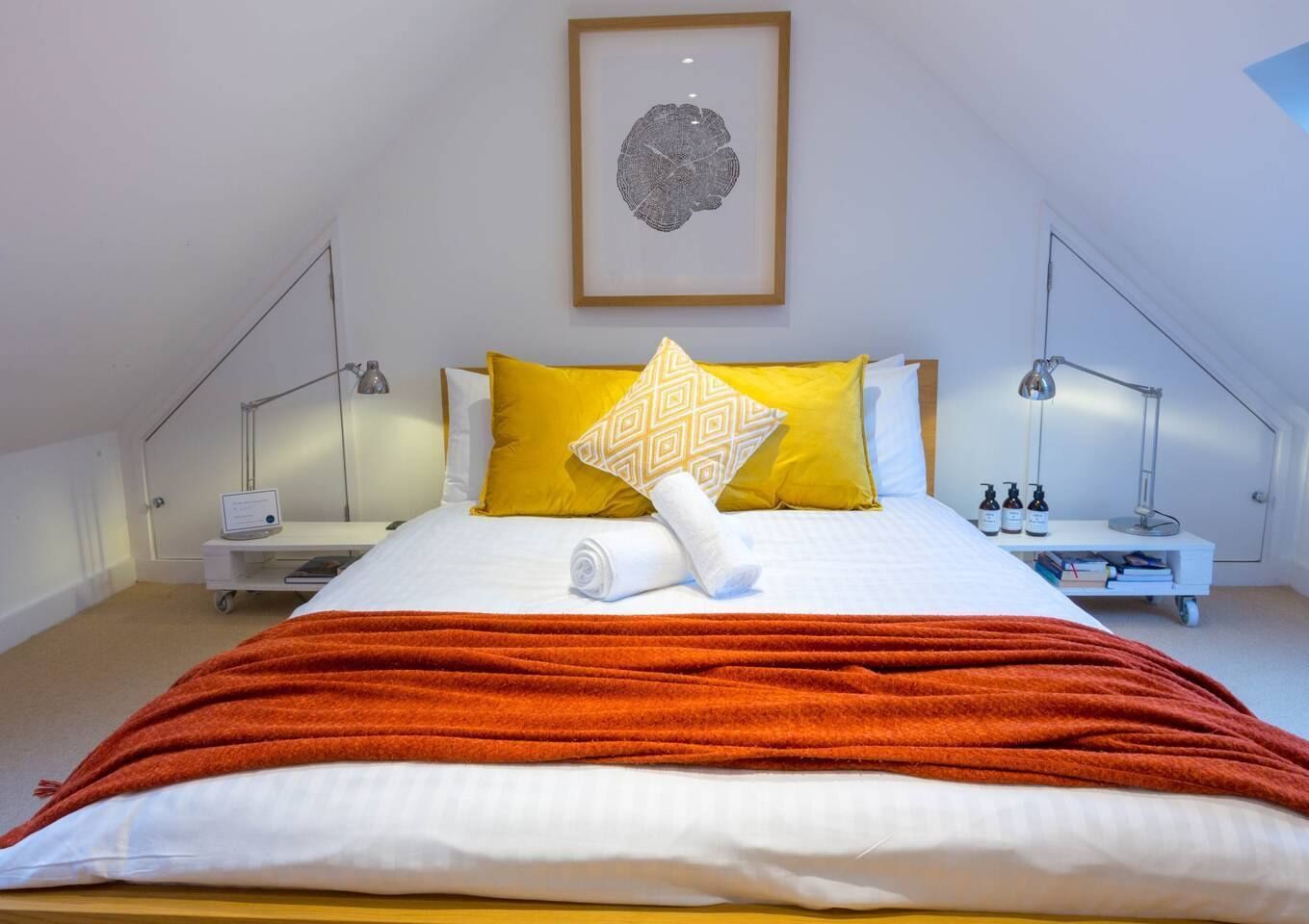 A neatly made bed with a white and orange blanket, two bedside tables with lamps, and a framed artwork on the wall.