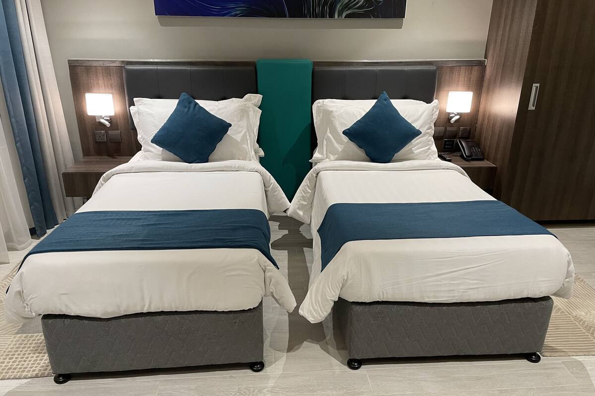 Habitación de hotel con dos camas, cada una con cubrecama blanco y azul, y un cojín decorativo verde. Se observan mesitas de noche con lámparas y un teléfono en un lado.
