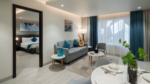 A modern hotel room with a grey sofa, a dining table set for two, a guitar, and a large window with curtains. - Citadines Al Ghubrah Muscat (Muscat)
