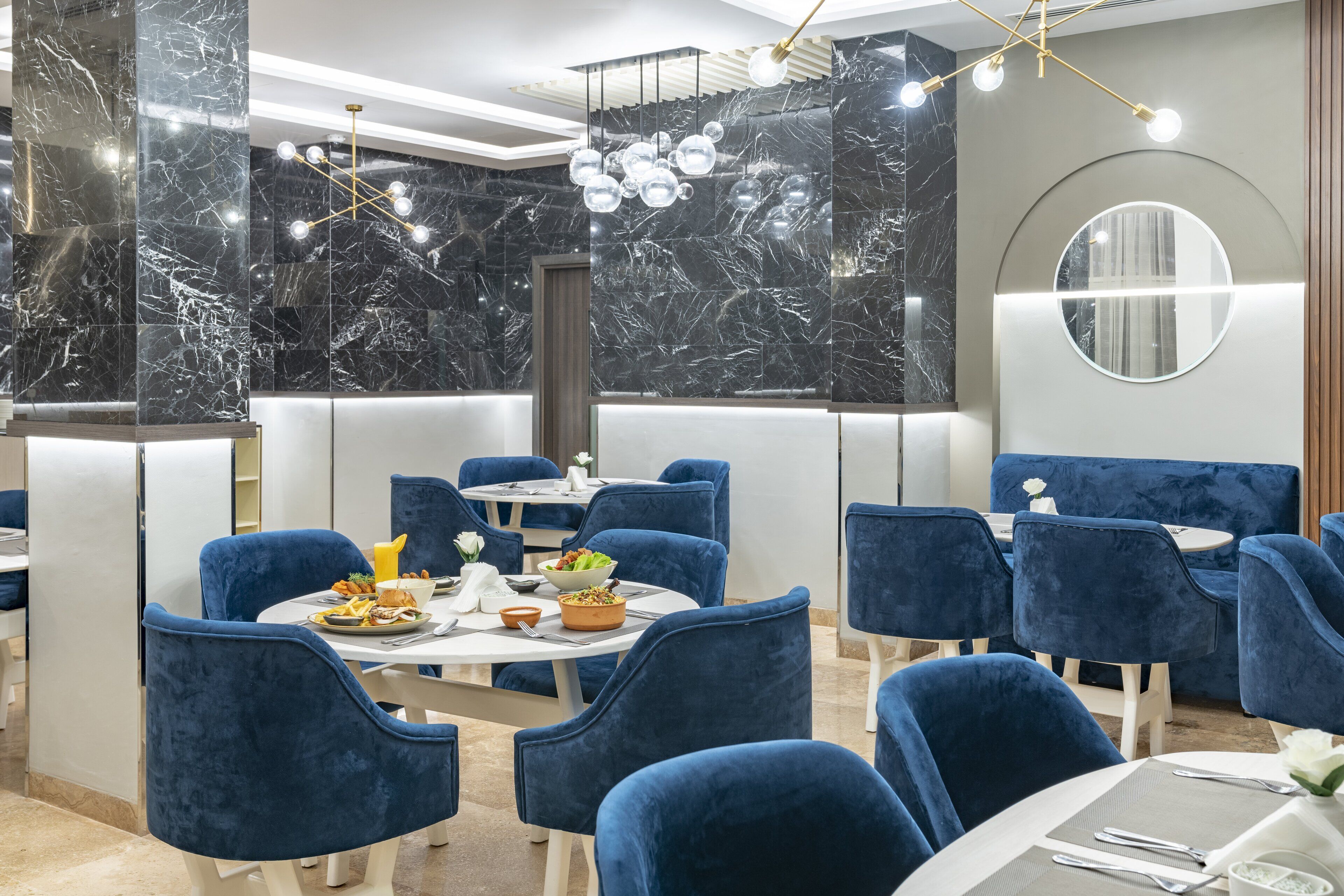 A modern dining area with marble walls, blue velvet chairs, and a chandelier.