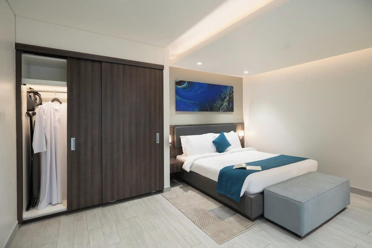 Un dormitorio moderno con una cama, un banco gris, un armario y un cuadro en la pared.