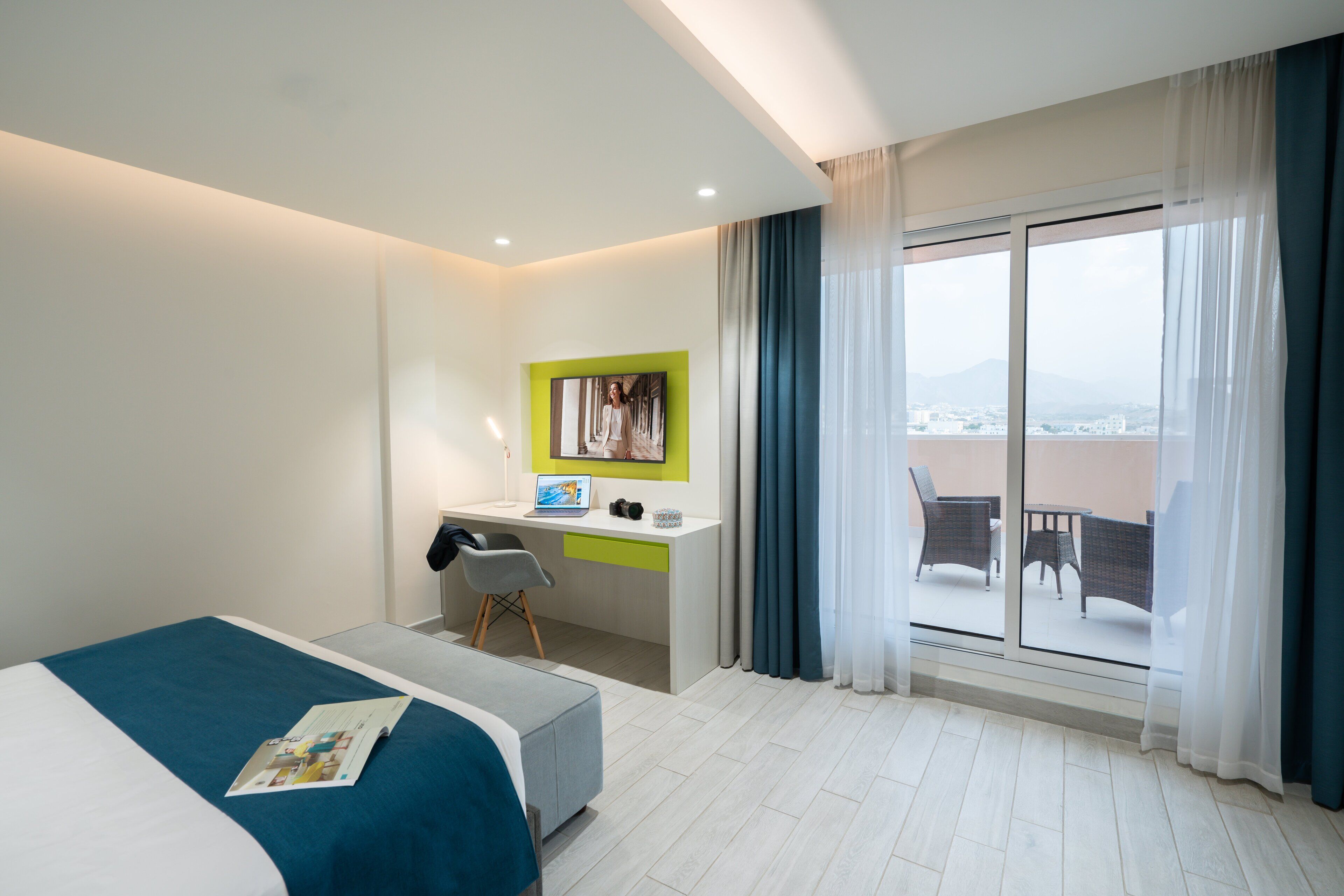 A hotel room with a bed, a desk, a chair, a window with curtains, and a balcony with furniture.