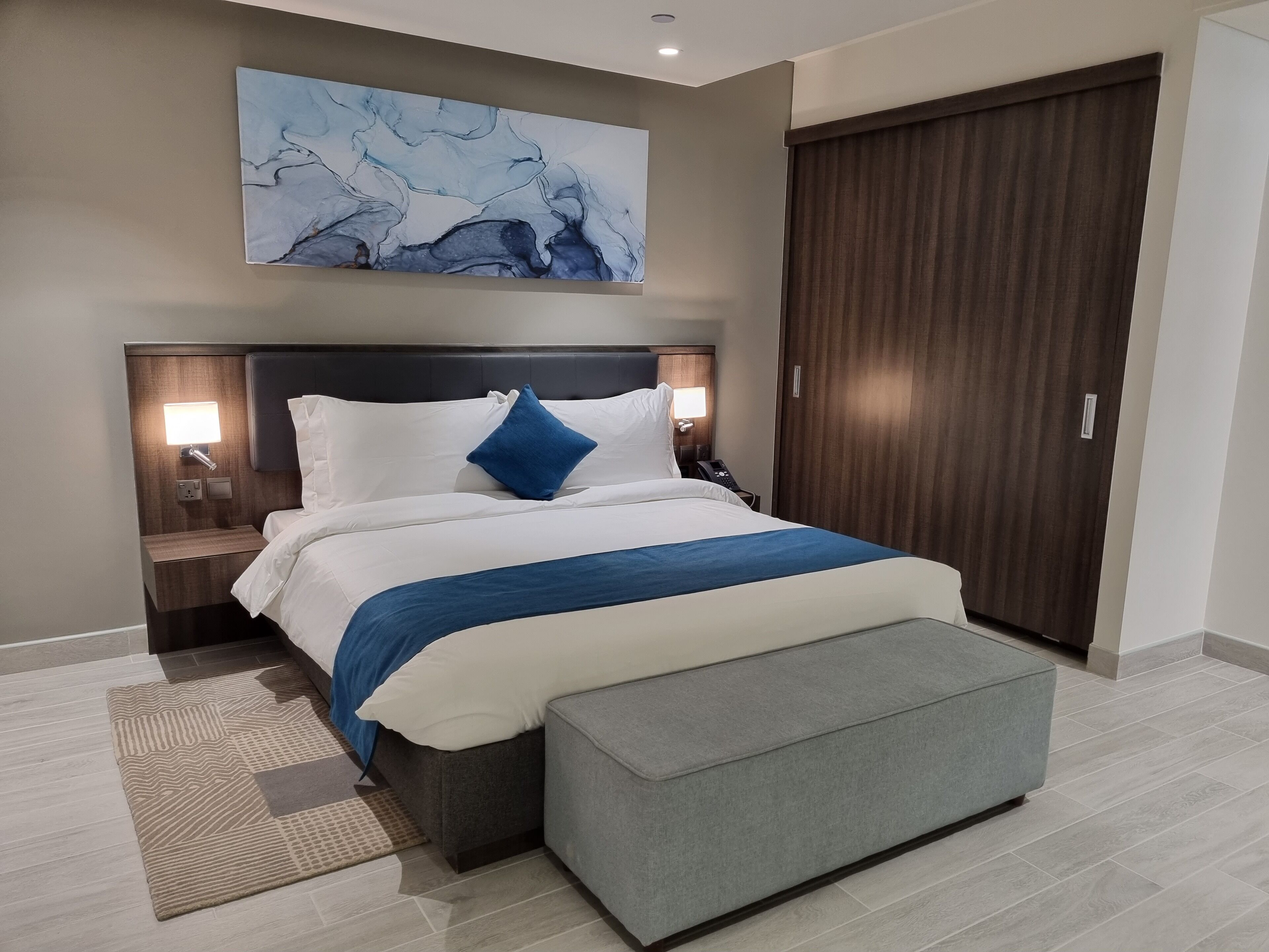 Ett modernt hotellrum med en stor säng, sänglampor, ett nattduksbord, en grå bänk, en trägarderob och en blå, abstrakt målning på väggen.
