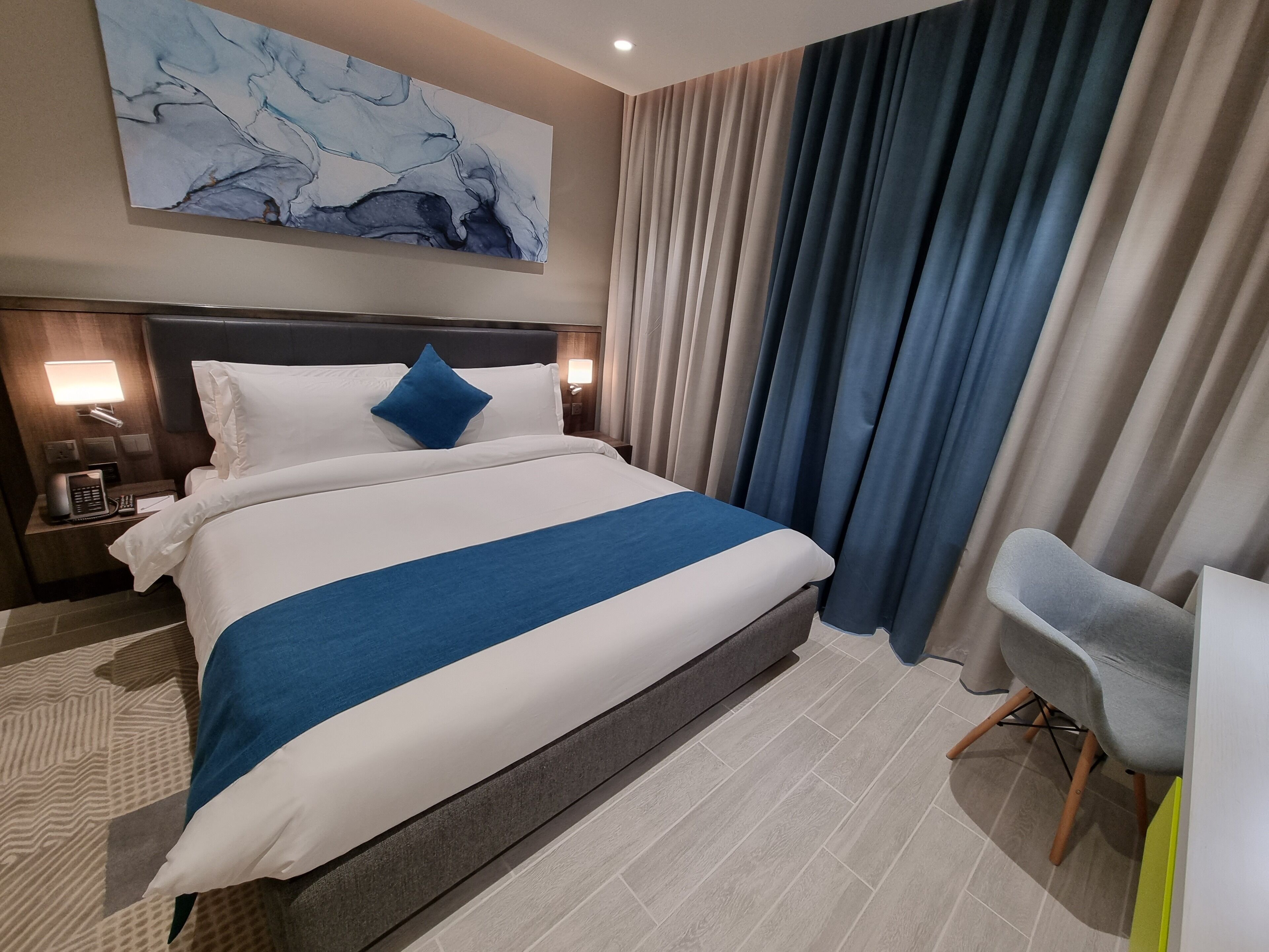 A hotel room with a large bed, a nightstand with a phone, a desk with a chair, and a painting on the wall.