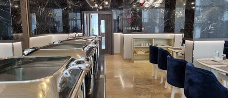 Restaurante moderno com paredes de mármore, um balcão comprido com bandejas de metal e cadeiras azuis.