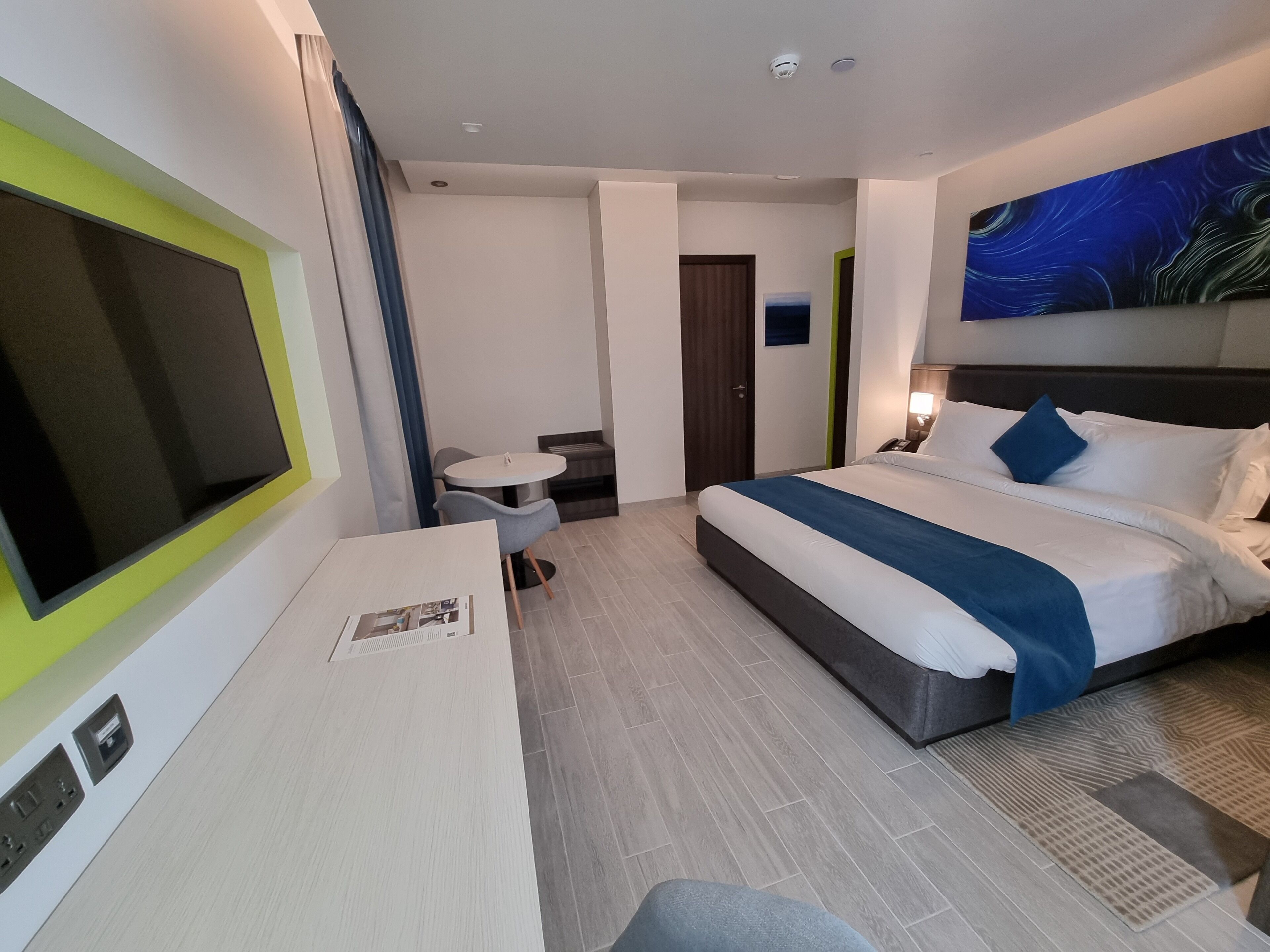 A modern hotel room with a bed, a desk, a chair, a small table, and a TV.