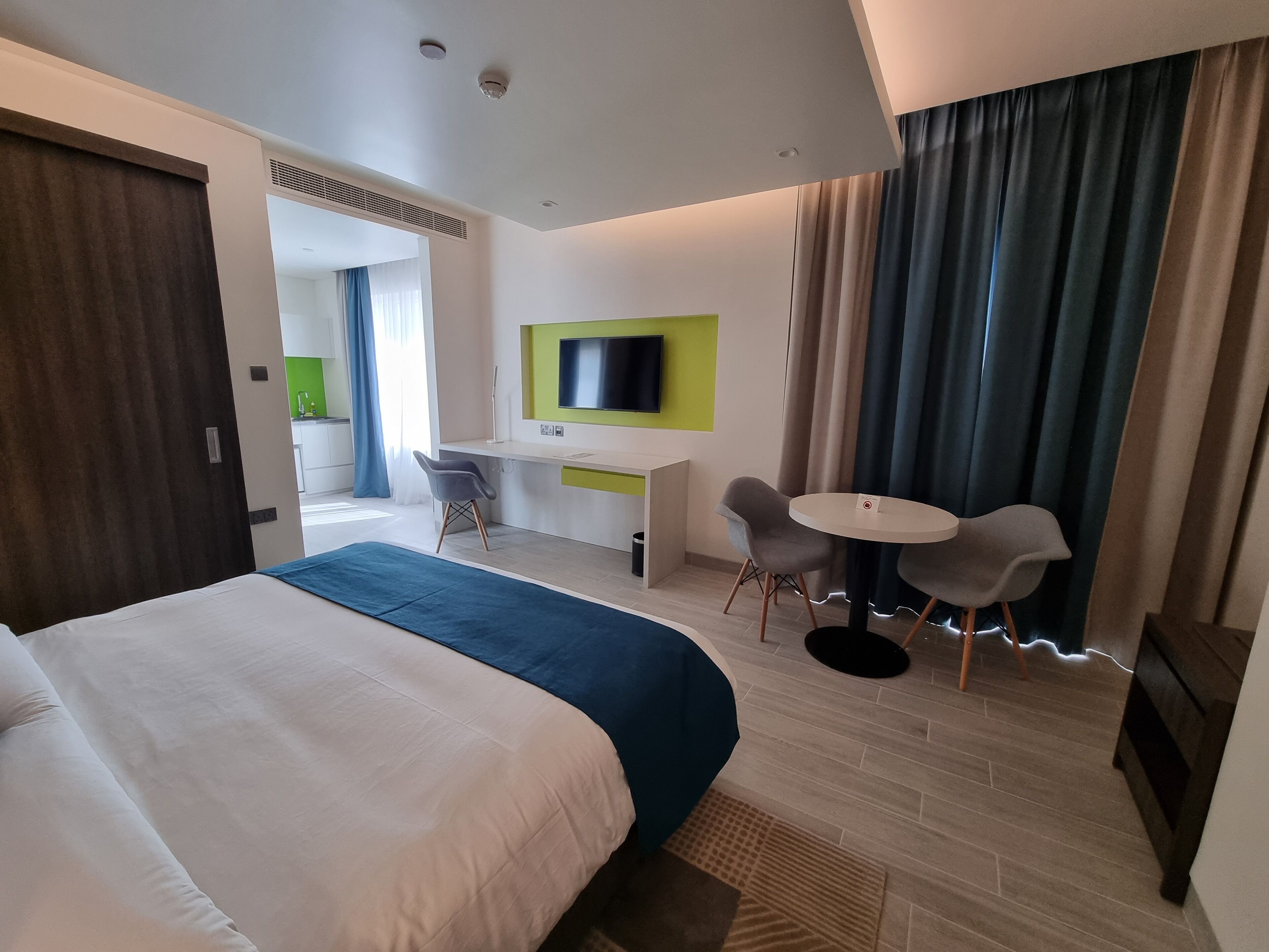 A hotel room with a large bed, a desk with a chair, a small round table, and a TV mounted on the wall.