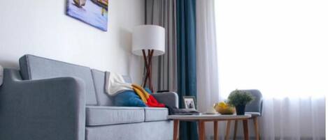 A modern living room with a grey sofa, a side table, a floor lamp, and a view of the outdoors through sheer curtains.