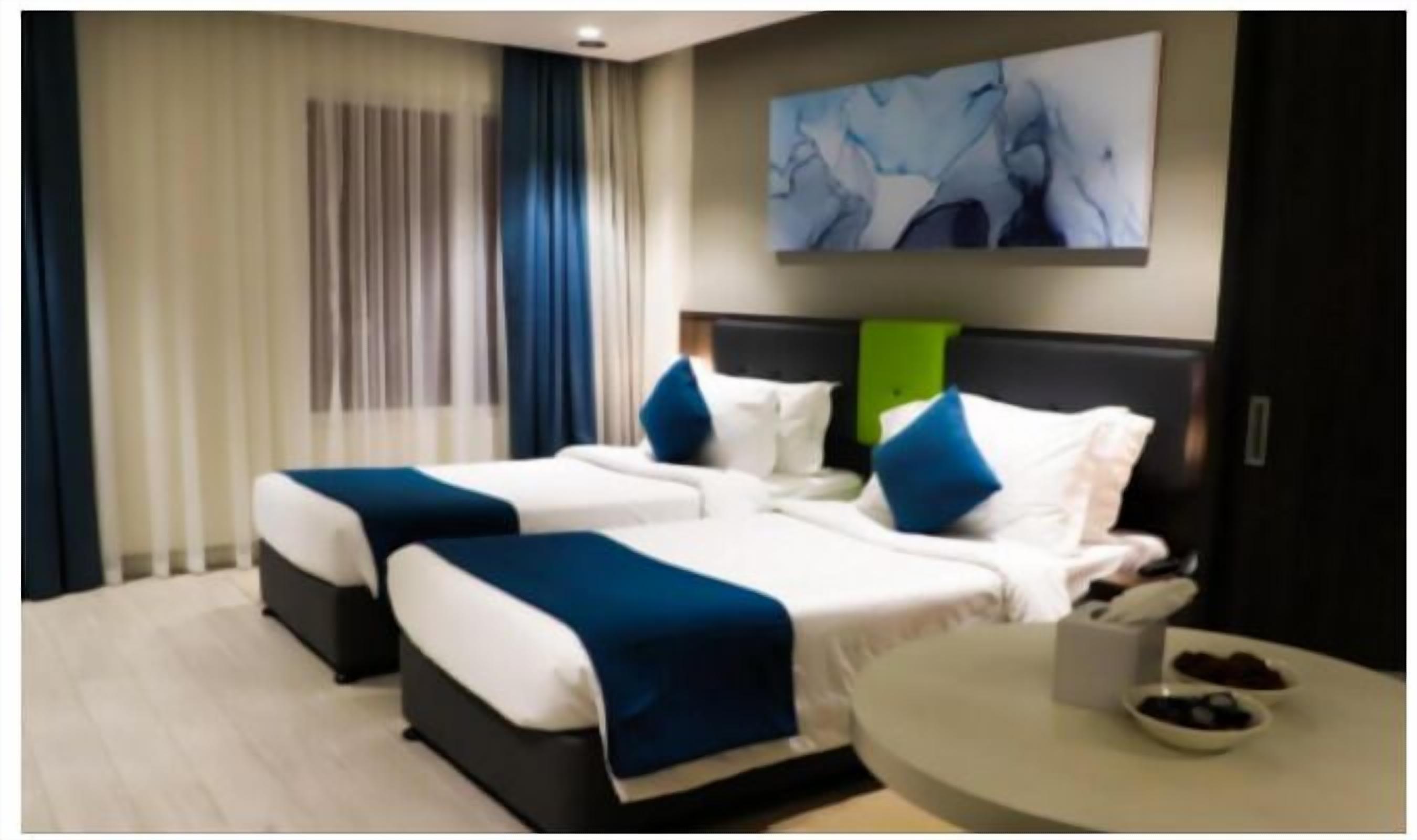 Une chambre d’hôtel avec deux lits, une table ronde sur laquelle sont disposés des bols de fruits, et un tableau accroché au mur.