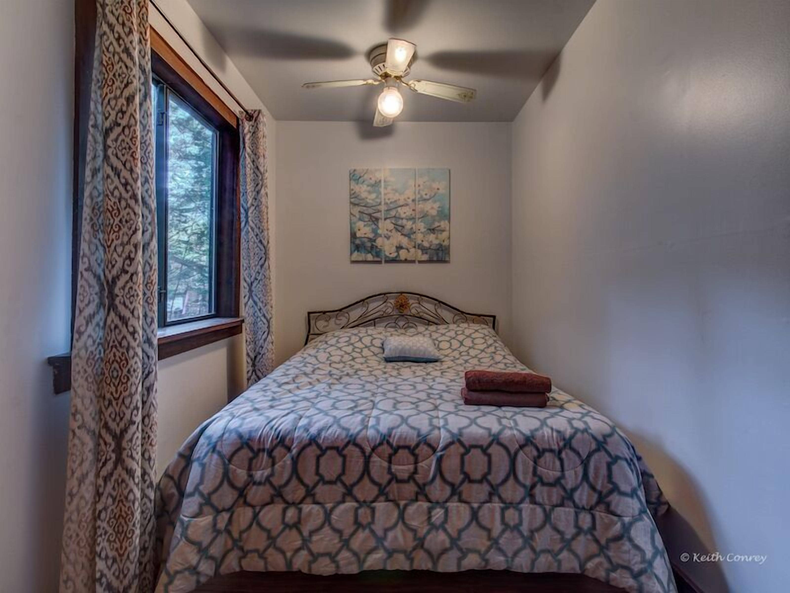 Un dormitorio con cama, ventilador de techo, ventana con cortinas y un cuadro en la pared.