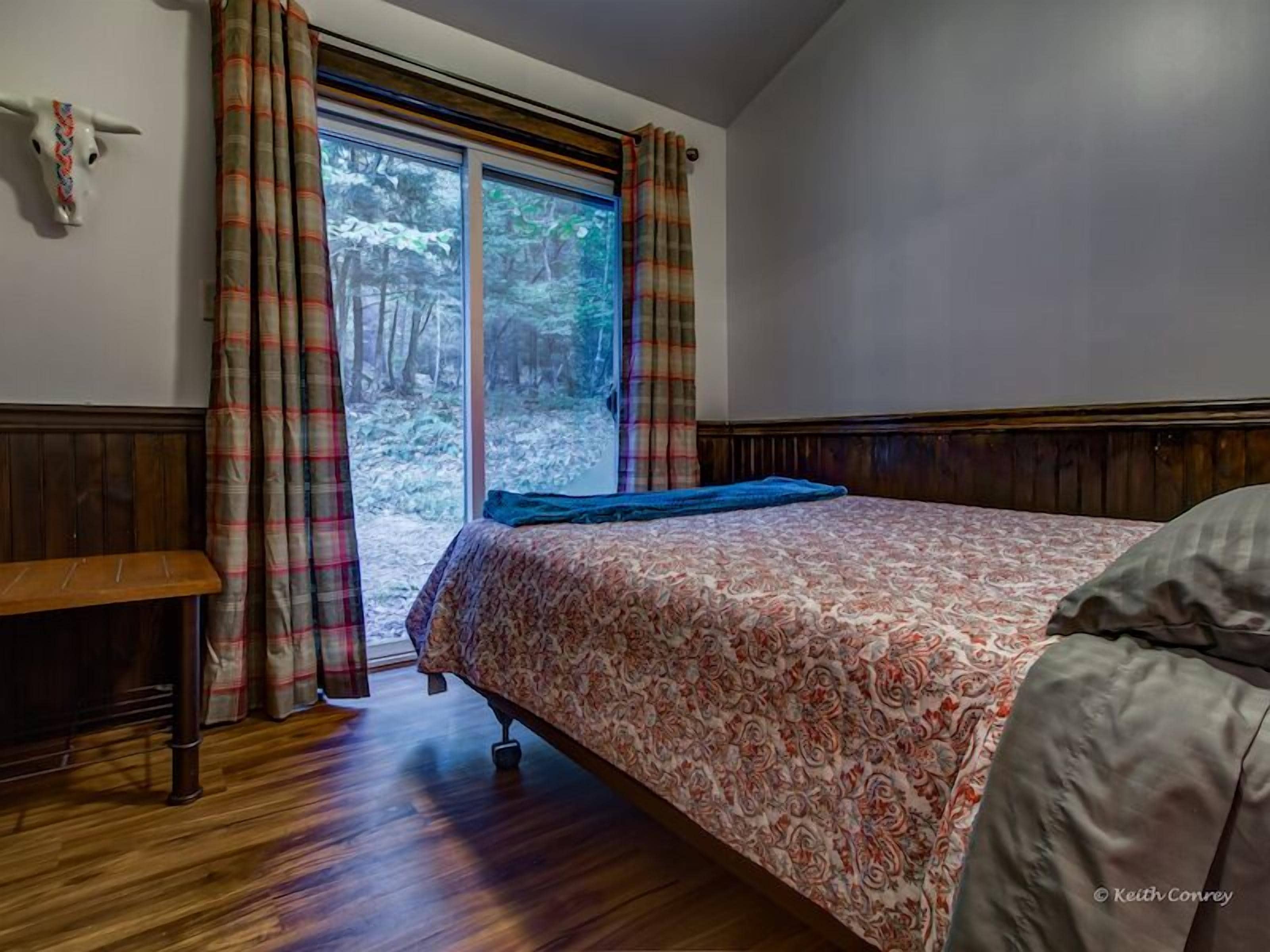 Een slaapkamer met een bed, houten vloer, geruite gordijnen en een raam met uitzicht op een bos.