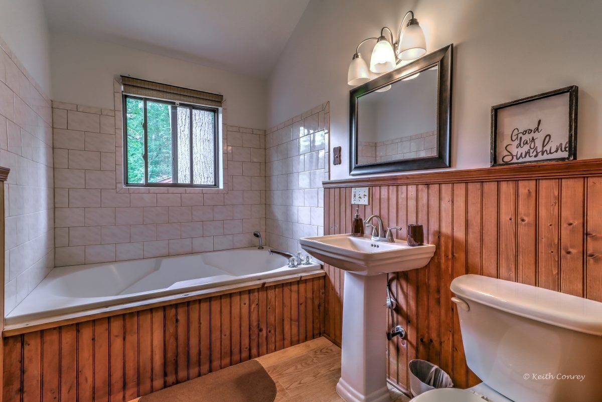 Ein Badezimmer mit Badewanne, Waschbecken und Toilette. Ein Fenster bietet Blick auf Bäume, es gibt einen Spiegel und ein Schild mit der Aufschrift 'Good day Sunshine'.
