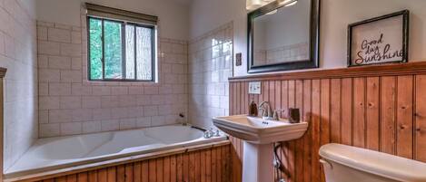Ein Badezimmer mit Badewanne, Waschbecken und Toilette. Ein Fenster bietet Blick auf Bäume, es gibt einen Spiegel und ein Schild mit der Aufschrift 'Good day Sunshine'.