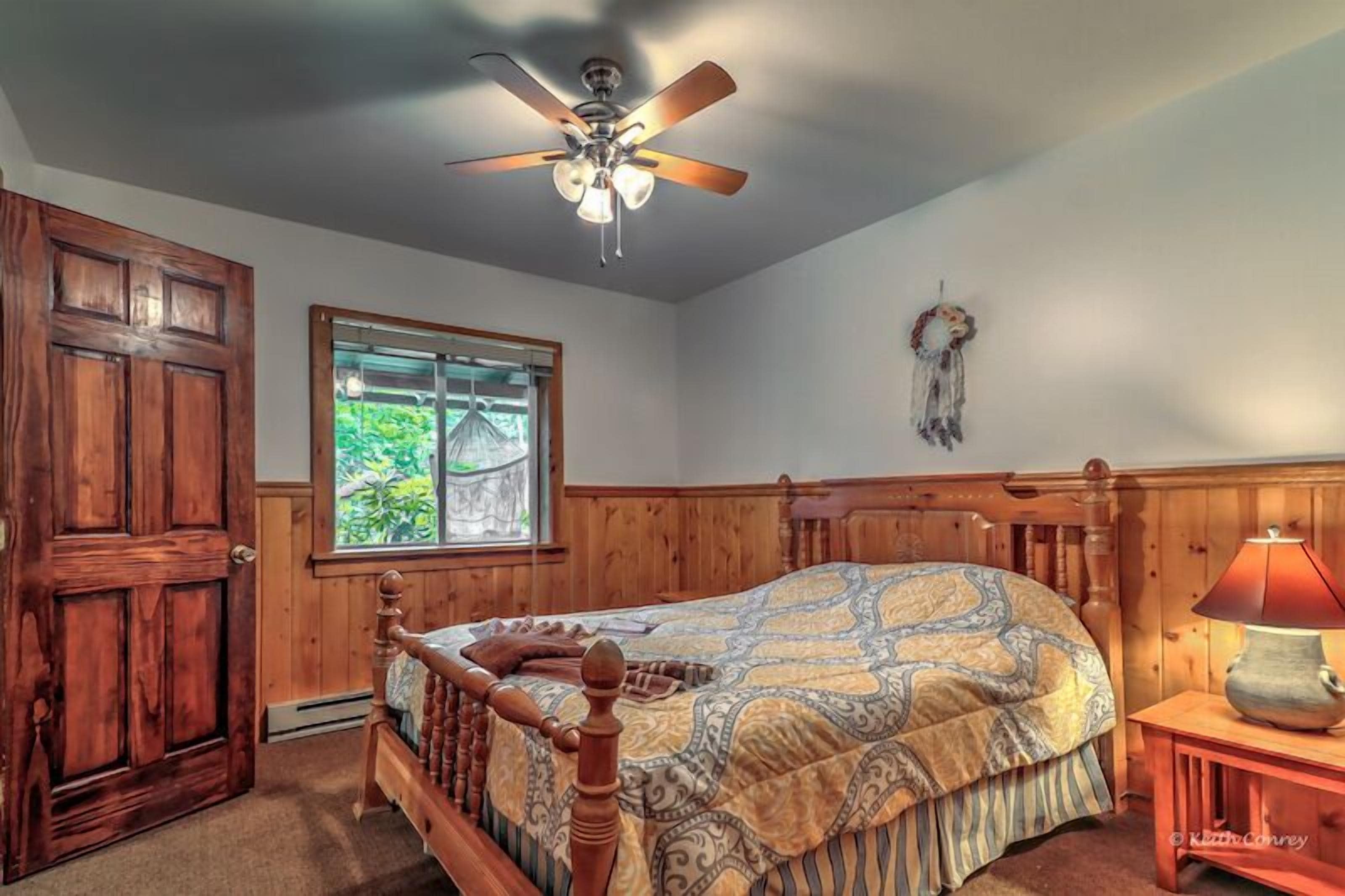 A bedroom with a wooden bed, a ceiling fan, a window with a view of greenery, and a dreamcatcher on the wall.