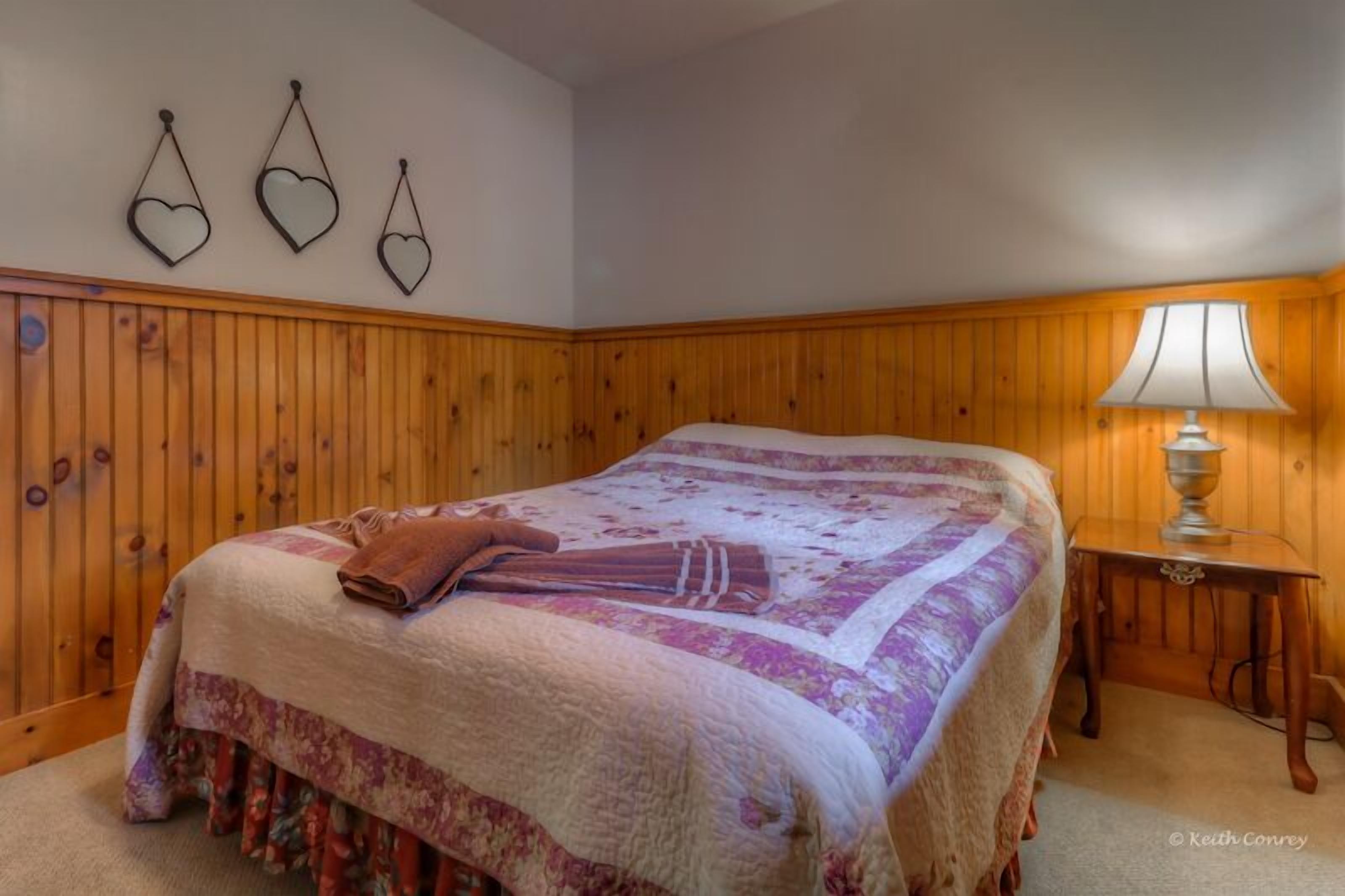 A bedroom with a large bed, wooden paneling on the walls, a bedside table with a lamp, and three heart-shaped wall decorations.