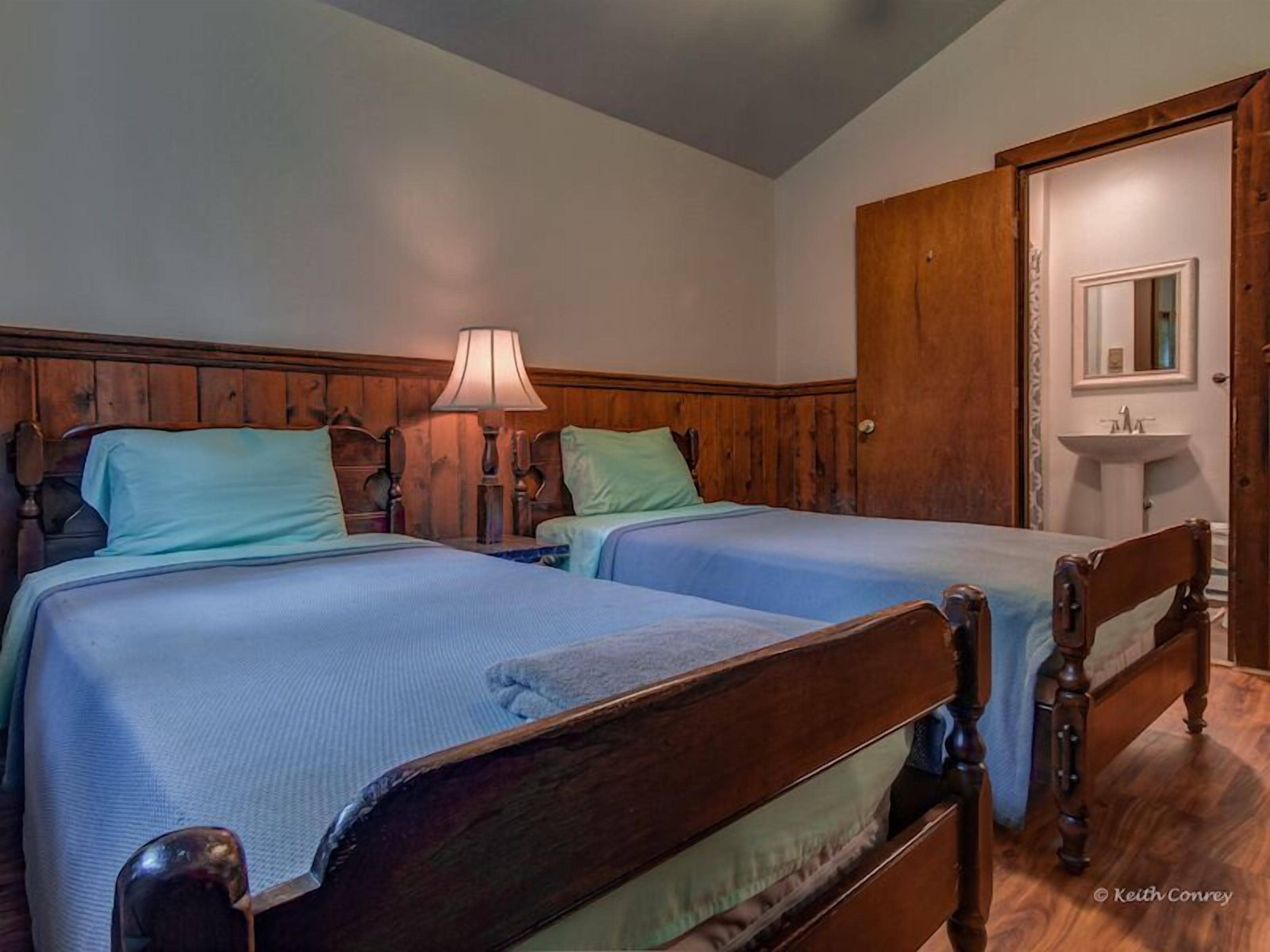 A room with two wooden beds, a wooden wardrobe, a bedside lamp, and a small sink.