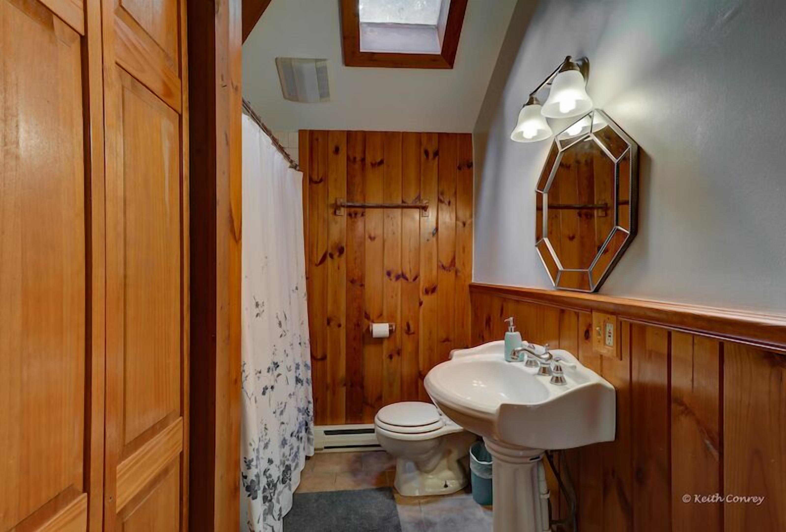 A bathroom with wooden walls, a white sink, and a skylight above the toilet.