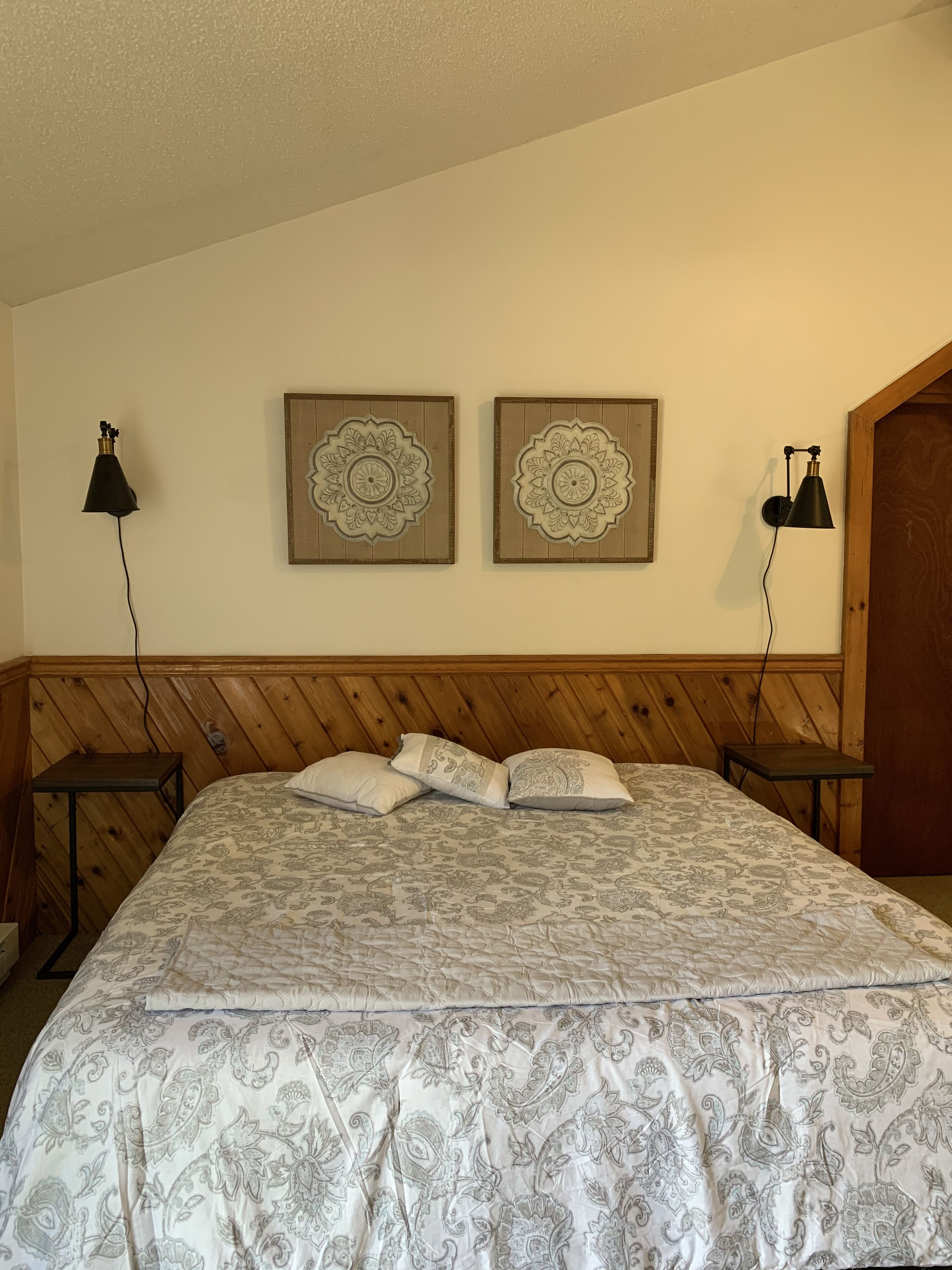 Une chambre à coucher avec une tête de lit en bois, un lit recouvert d’une literie à motifs, deux lampes fixées au mur et deux éléments décoratifs muraux.
