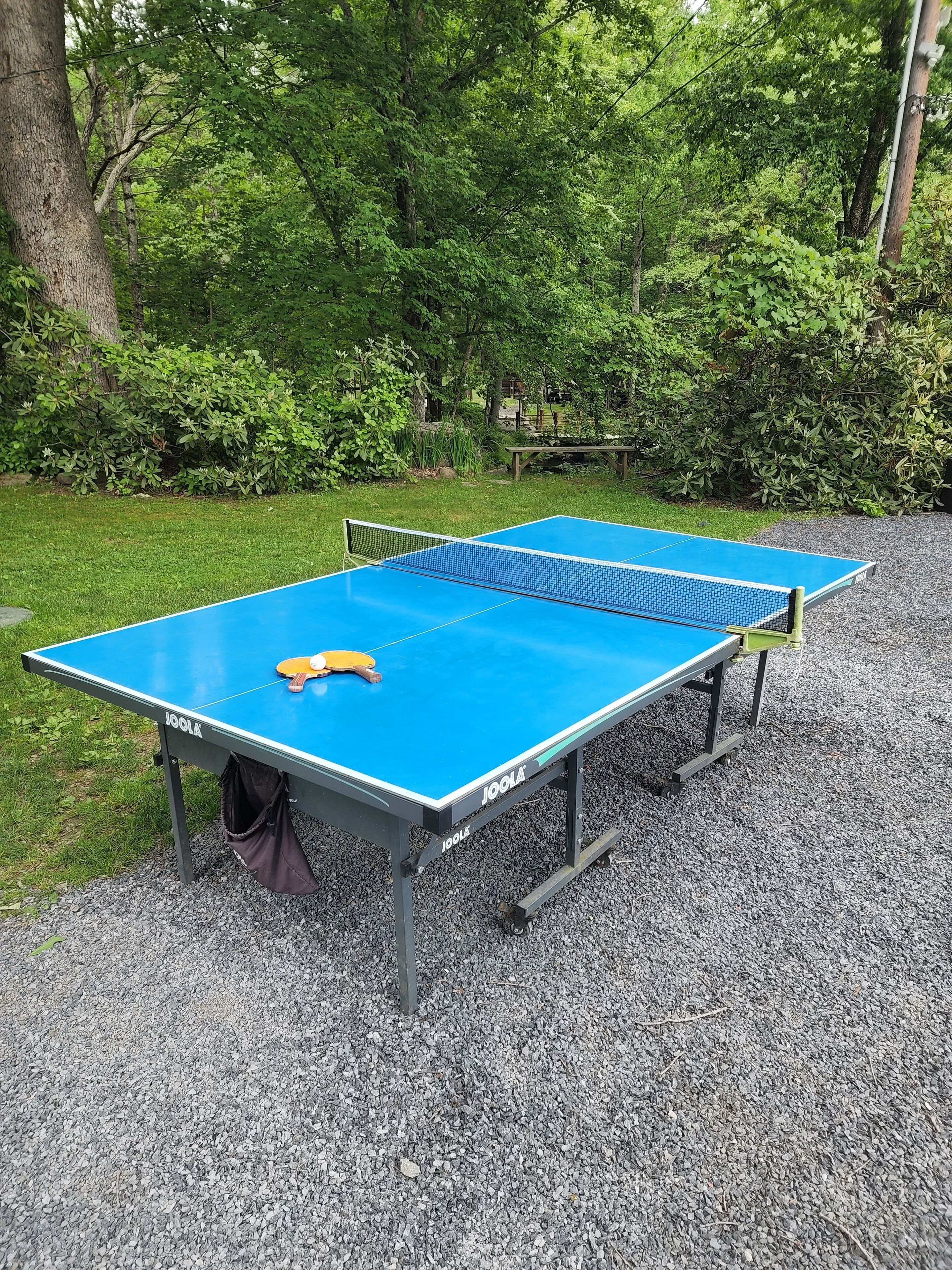An outdoor ping pong table with a net, surrounded by gravel and greenery.