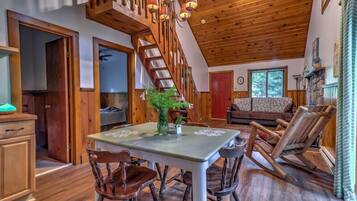A cozy cabin interior with a dining table, chairs, a staircase, and a living area with a couch and a rocking chair.