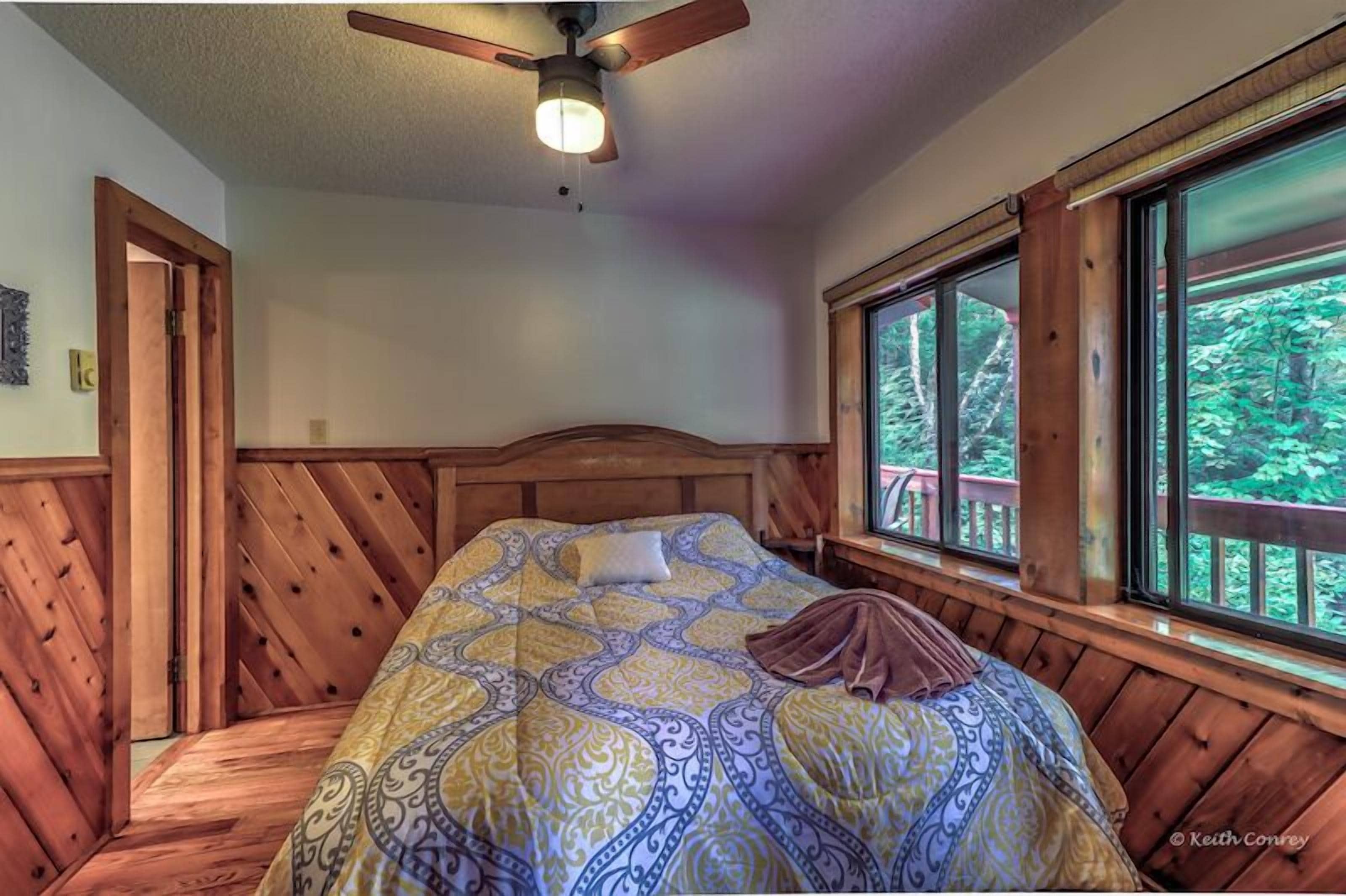 A wooden bed with a patterned bedspread, a ceiling fan, and a window with a view of trees.