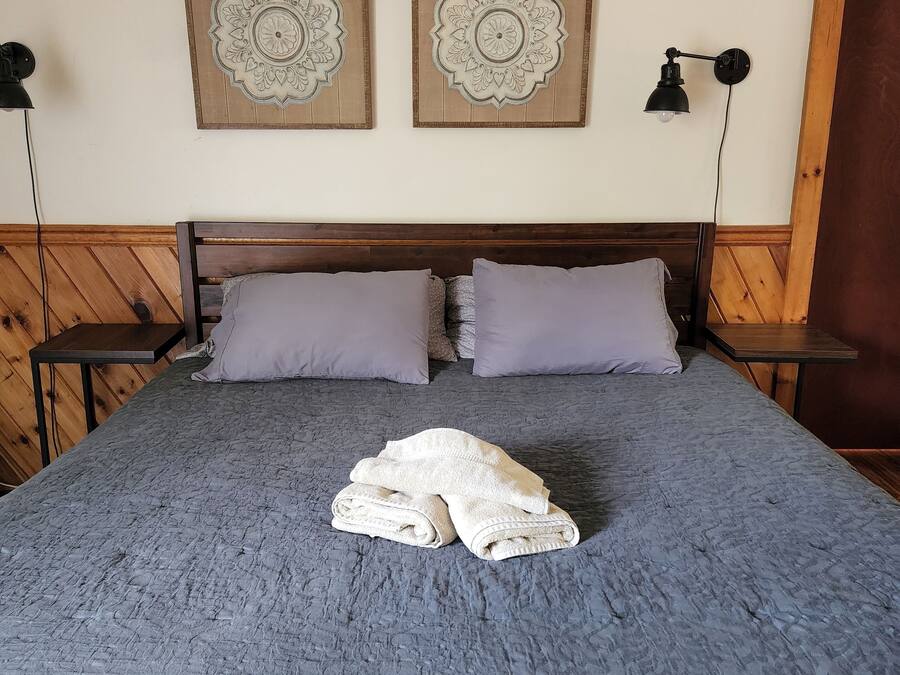 Una cama con una manta gris texturizada, dos almohadas y una toalla blanca doblada. Encima de la cama, hay dos paneles decorativos montados en la pared.