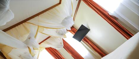 A room with a bed covered in white and yellow drapes, a black object on the wall, and red curtains.