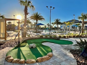 A golf putting green surrounded by palm trees and lounge chairs, with a pool and umbrellas in the background.