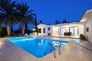 A swimming pool with a clear blue water surface, surrounded by a tiled deck. There are lounge chairs and a dining area with tables and chairs. Palm trees are visible in the background. - Luxurious, Spacious, FULLY air conditioned villa set in a serene environment! (Paphos)