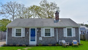 Une maison de plain-pied avec un toit recouvert de bardeaux gris, une cheminée en briques et une porte d’entrée bleue. On peut voir deux chaises Adirondack dans le jardin.