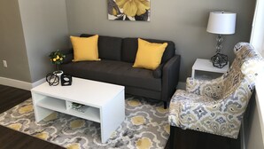 Een woonkamer met een grijze bank, twee gele kussens, een witte salontafel, een gestreept fauteuil, een vloerlamp en een groot bloemenportret aan de muur.