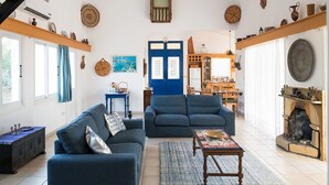 Een woonkamer met twee banken, een salontafel en een open haard. Er staan planken met decoratieve objecten en in de achtergrond is een eethoek te zien.