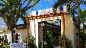 Une entrée sécurisée, fermée par un portail, mène à la résidence TAMARINDO BLUE APARTMENTS, agrémentée d’une pergola en bois et de palmiers.