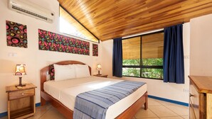 Une chambre avec un plafond en bois, un lit recouvert d’une couverture bleue, deux tables de chevet avec des lampes et une fenêtre avec des rideaux.