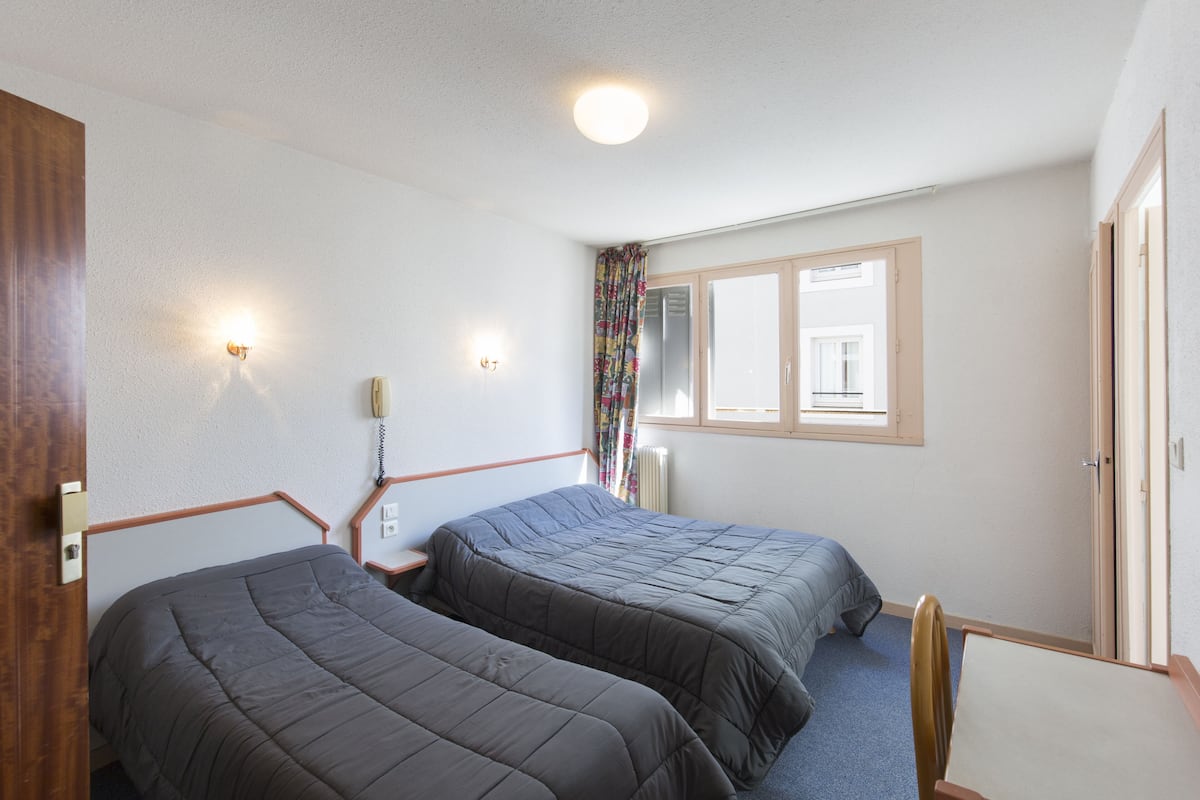 A room with two beds, a desk, and a chair. There is a window with curtains, a radiator, and a wall-mounted light fixture.