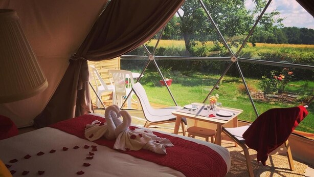 A cozy tented room with a bed adorned with rose petals and a swan-shaped towel arrangement. A table set for two is positioned near a window overlooking a lush garden. - Tui's Nest Glamping (Villentrois-Faverolles-en-Berry)