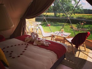 A cozy tented room with a bed adorned with rose petals and a swan-shaped towel arrangement. A table set for two is positioned near a window overlooking a lush garden. - Tui's Nest Glamping (Villentrois-Faverolles-en-Berry)