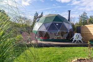 A geodesic dome structure with a chimney, surrounded by a wooden deck, chairs, and a grassy area. - Tui's Nest Glamping (Villentrois-Faverolles-en-Berry)