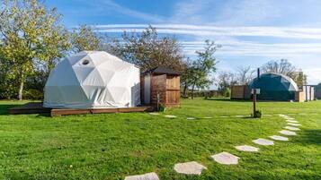 A white geodesic dome tent set on a grassy lawn with a wooden structure nearby.