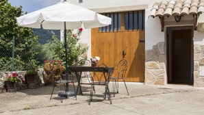 Un patio al aire libre con una mesa, sillas, un paraguas blanco, plantas en macetas y una puerta de madera.