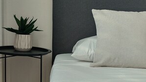 Una cama bien tendida con cabecera gris, una mesita redonda con planta en maceta y una pared lisa.