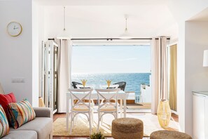Un salon moderne doté d’une grande porte-fenêtre coulissante donnant sur un balcon avec vue sur la mer. On y trouve un canapé blanc agrémenté de coussins colorés, une table à manger avec des chaises et un grand vase.