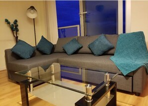 A modern living room with a grey sectional sofa, teal cushions, a glass coffee table, and a floor lamp. - 47 Chelsea House - Q-zotica City Centre Apartments: Parking, City view & Smart gadgets (Milton Keynes)