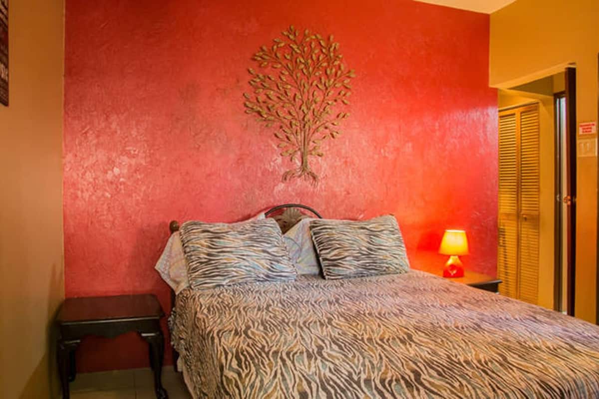 Un dormitorio con una colcha de estampado de cebra, un cabecero de madera, una mesita de noche con lámpara y una pared roja con un mural de un árbol.