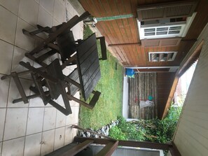 A patio with a tiled floor, a wooden table and chairs, a blue water container, and a small garden area with a wooden fence and some greenery.